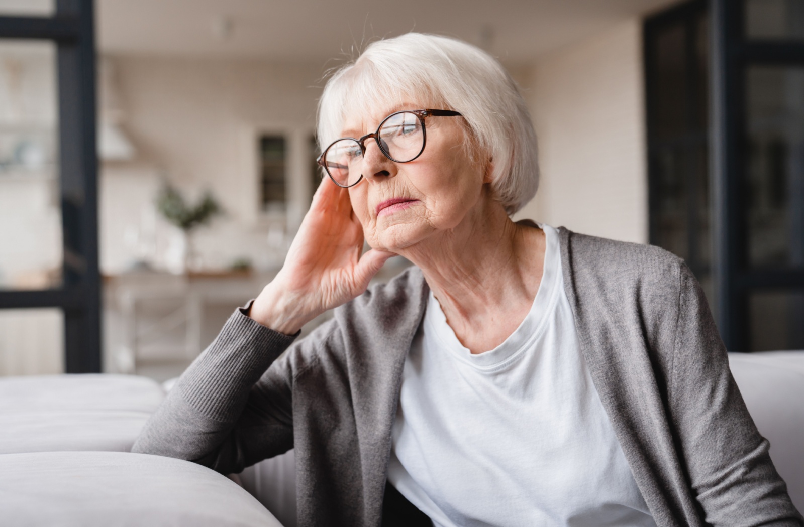 A senior woman with glasses longingly looks out of a window