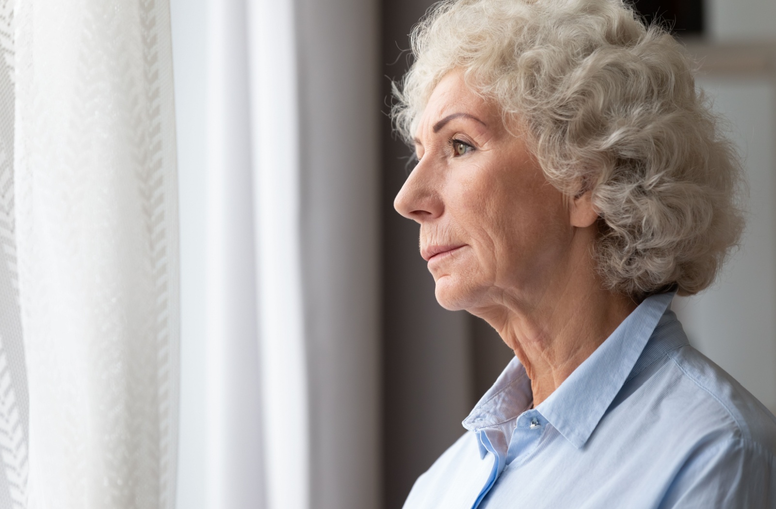 A senior woman looking out the window at a Senior Living Community looking thoughtful.