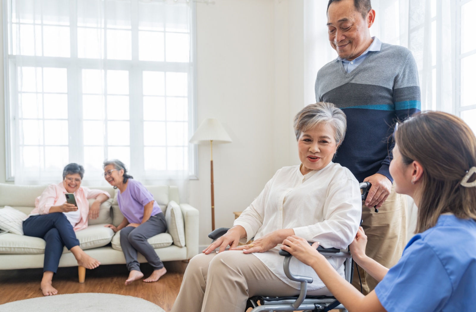 A senior woman being dropped off at a senior living community by her son for respite care with other seniors in the background enjoying themselves.