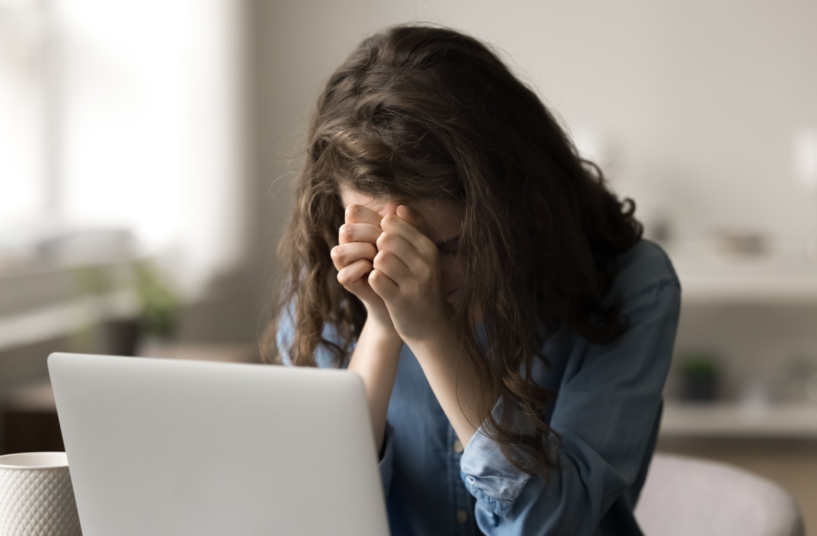 A young adult with their head in their hands from dealing with severe caregiver fatigue, researching nearby senior living communities on their laptop.