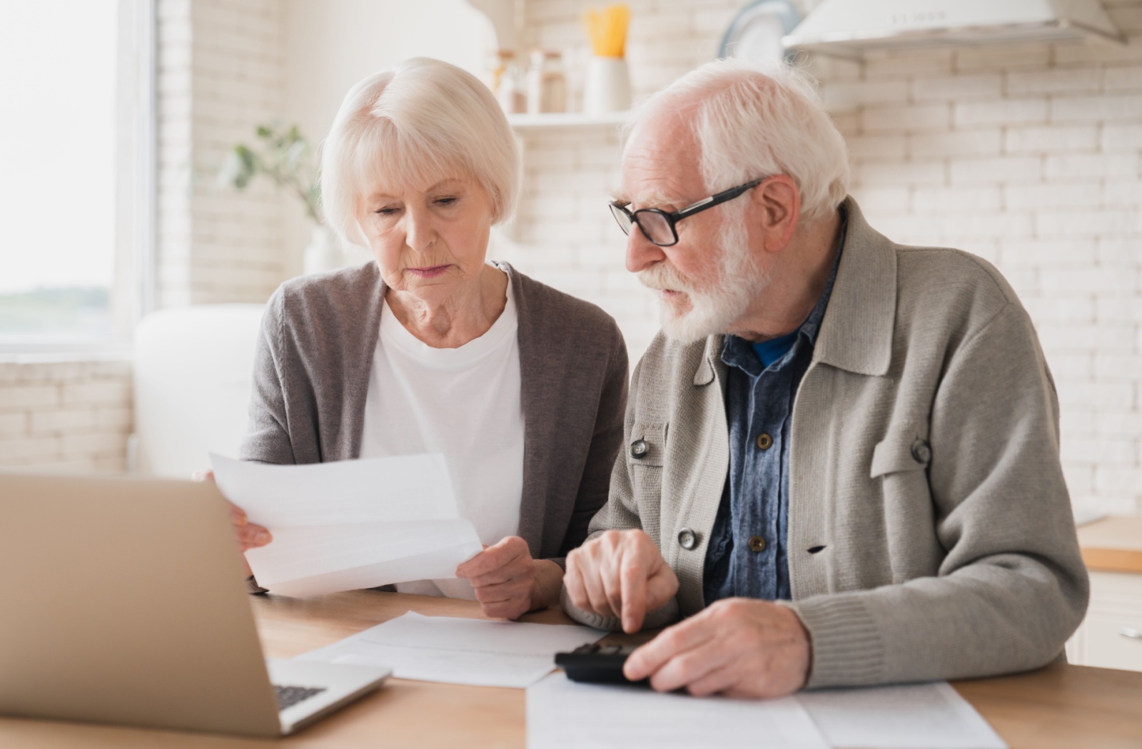 Senior woman and man looking at papers in front of a laptop. The man is using a calculator.