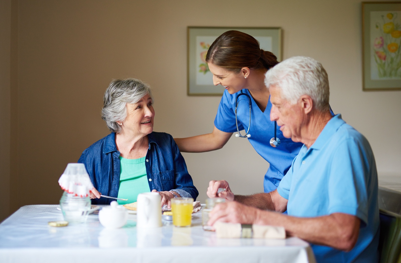 Nurse in blue scrubs greeting senior man and woman eating breakfast at a table.
