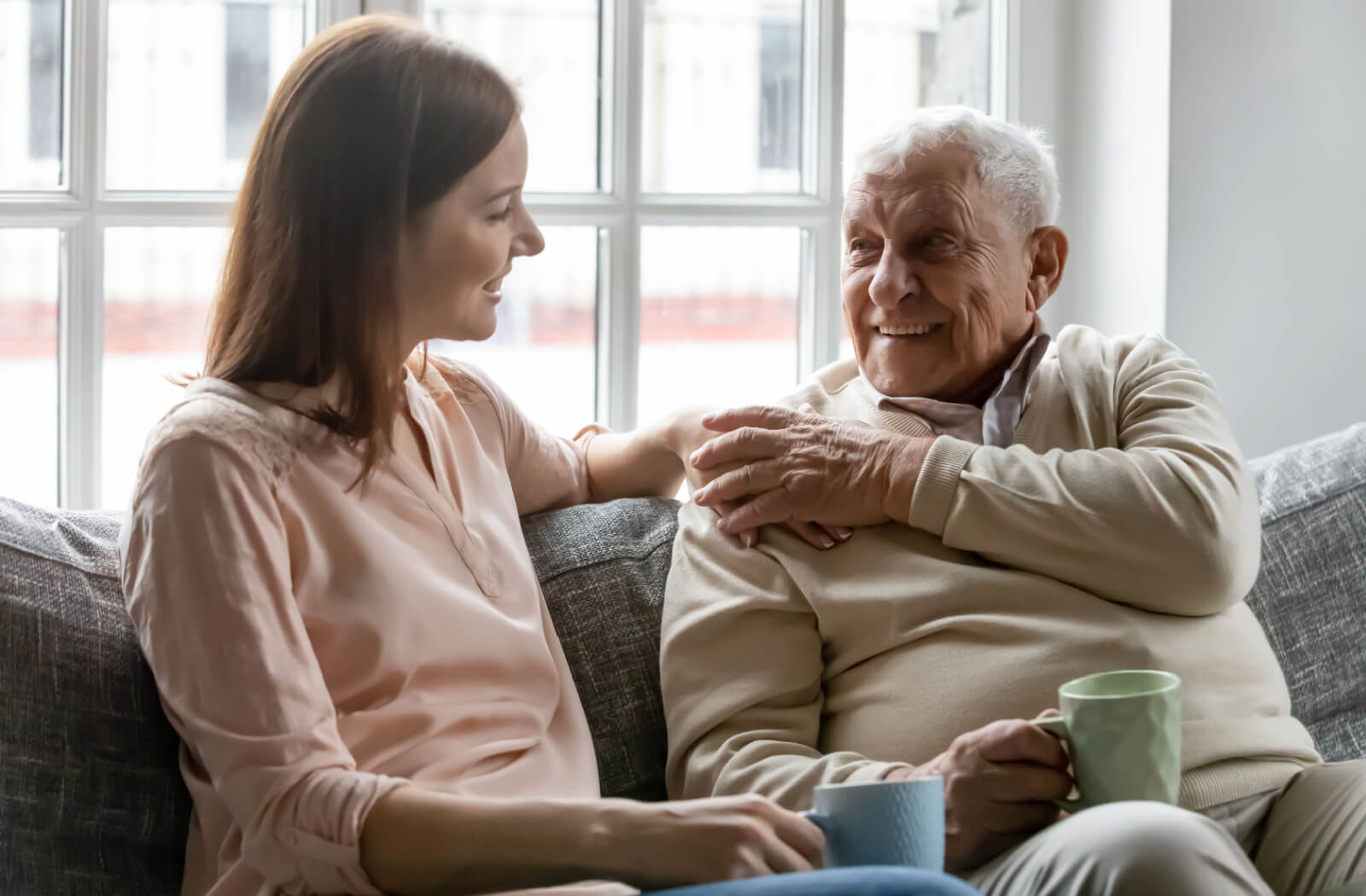 An adult child visiting their older parent in memory care, sitting on the couch and drinking coffee together.