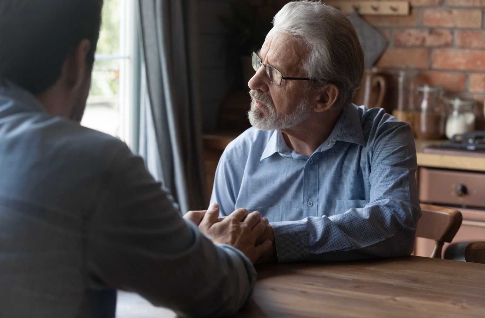 An adult child sitting across a kitchen table from their older parent, holding their hand to comfort them.
