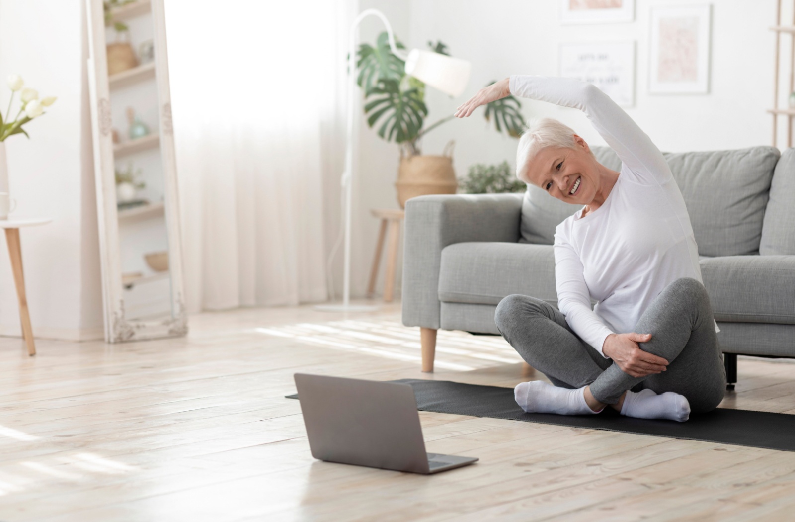An older adult sits cross-legged and stretches an arm overhead while following an online hip exercise tutorial