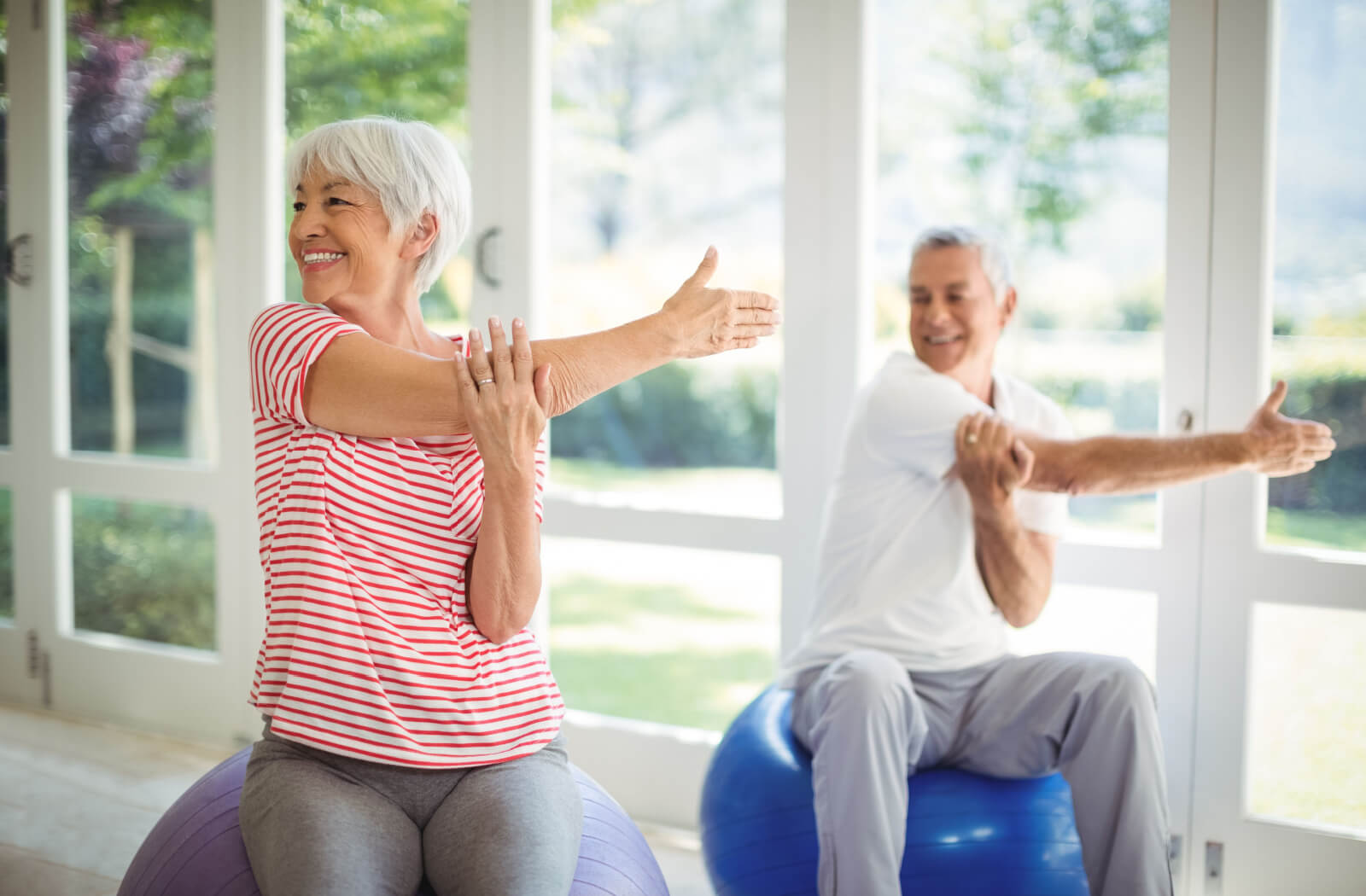 2 older adults sitting on medicine balls stretching their arms and twisting to strengthen their hips
