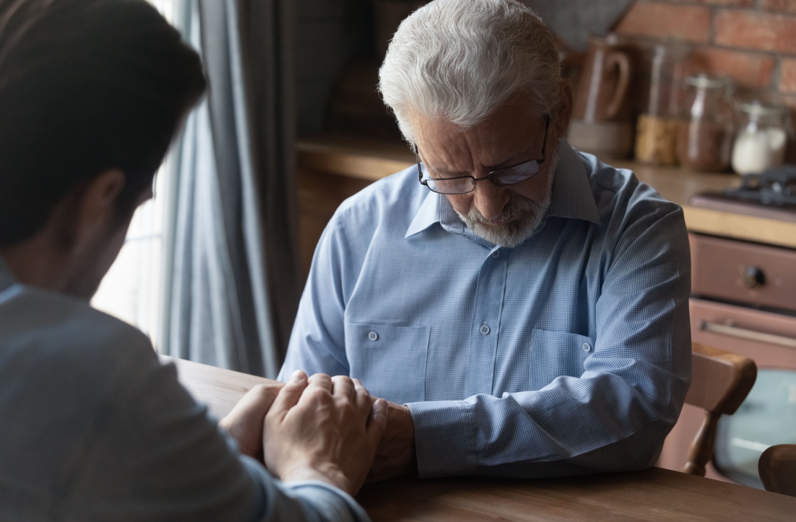 An adult holds their older parent’s hand for emotional support as they talk about progressing memory loss