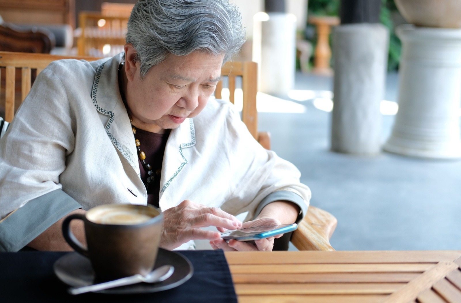 A senior scrolls on a smartphone while enjoying a cup of coffee outside at a wooden table.
